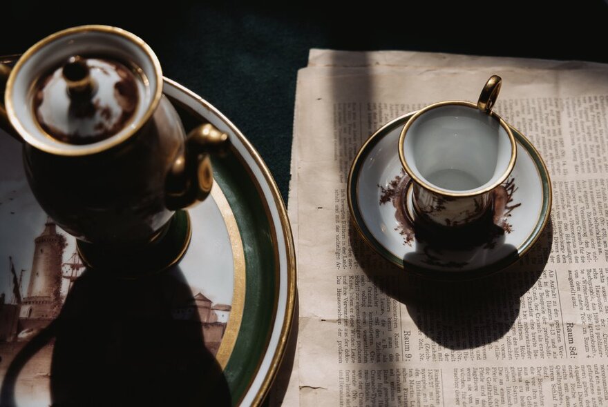 A Regency-era tea cup and saucer sitting on an old-fashioned newspaper, with a painted tray and teapot next to it.