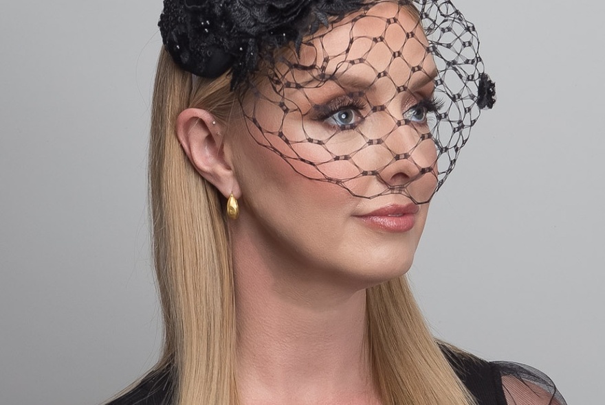 A headshot of a woman wearing a black fascinator with netting over her face. 