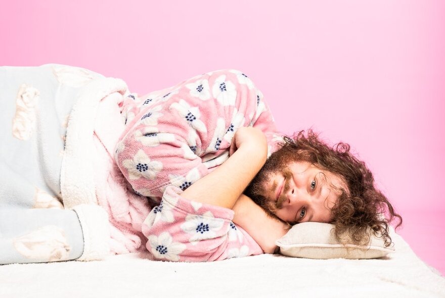 Man wearing a dressing gown, lying on his side covered with a blanket and looking cosy and snug.