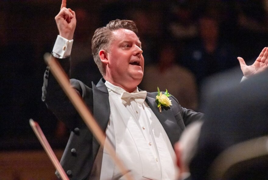 A conductor wearing a white shirt and black jacket, with his arms outstretched, holding a baton.