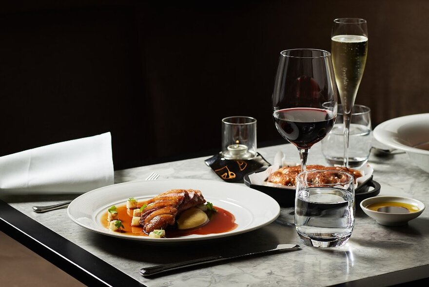 Table set with a meat dish in a white bowl with a glass of red wine, flute of bubbles and glasses of water.