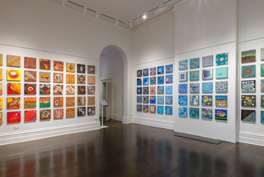 An installation view of many different paintings of the same size (30x30cm) displayed on two walls in a gallery. 