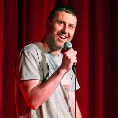 Comedian Lloyd Langford holding a microphone in his hand and performing stand-up comedy on a stage against a red curtain.