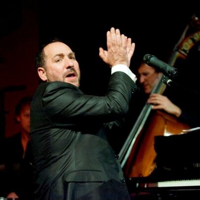 Steve Sedergreen clapping his hands on stage in front of a piano with a double bass player in the background. 