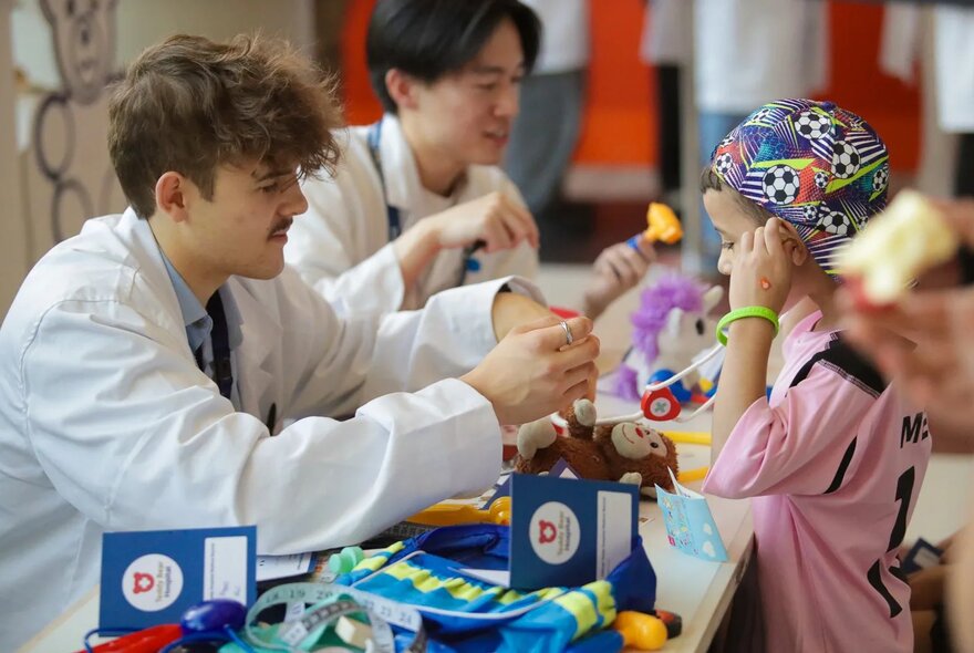 Adults wearing white doctor's coats, seated at a low table interacting with children and teddy bears, at the teddy bear hospital at the Kids Day Out.