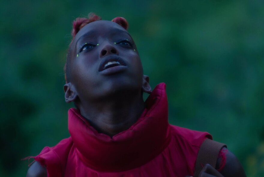 A still from the film Neptune Frost, of a young person looking up, wearing an outdoor jacket, blurred greenery behind them.