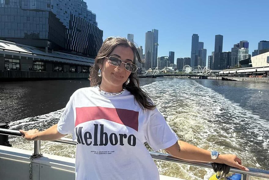 A woman wearing sunglasses and a t-shirt posing on a cruise boat, with a view of the Melbourne skyline and Yarra river behind her.