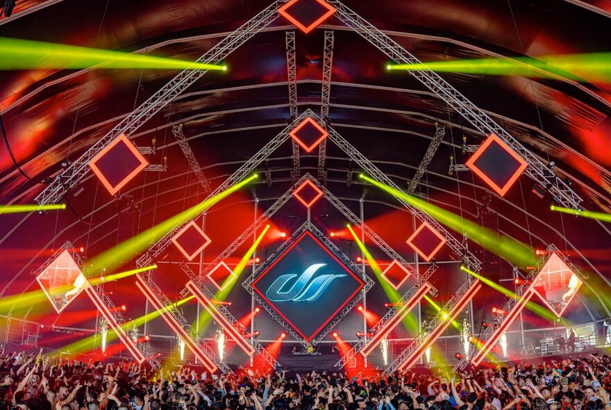 A scene from a trance music festival with a huge crowd and neon projections and lights above them, most in diamond shapes and yellow beams. 