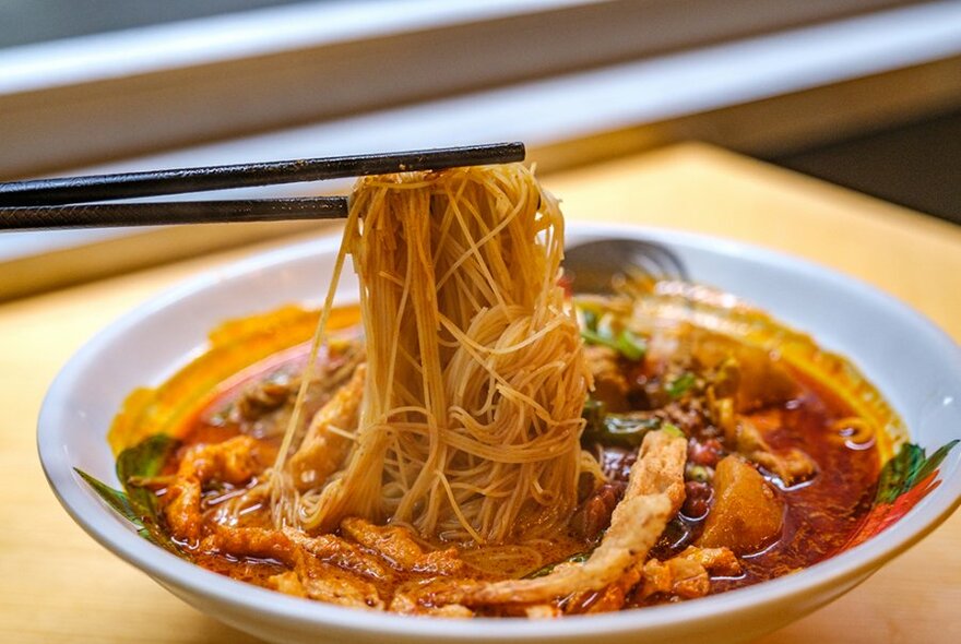 A pair of chopsticks lifting noodles out of a bowl of soup.