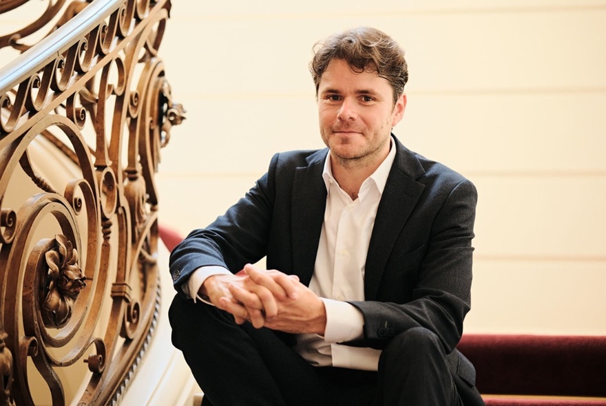 Portrait of conductor, Nicholas Carter, sitting on a velvet covered staircase with an elaborate balustrade.