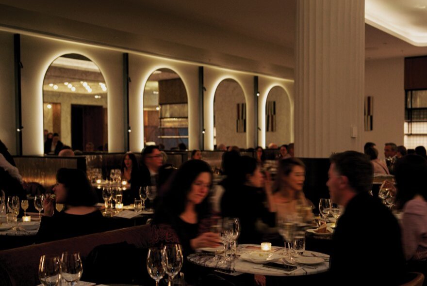 The dimly lit interior of a restaurant, with patrons seated at marble tables and booths in front of arched mirrors.