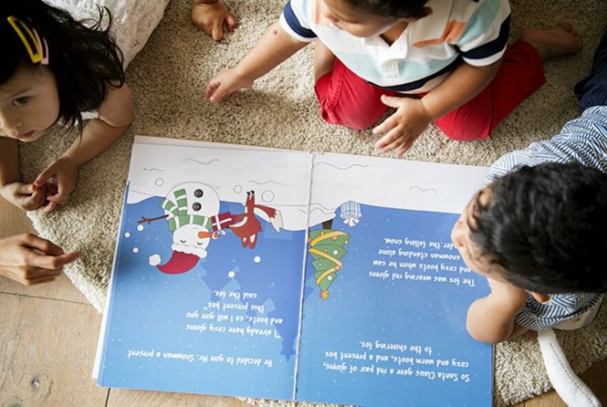 Looking down on three children on a floor crowded around an open picture book about Santa. 