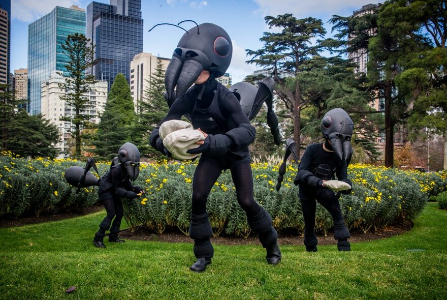 Performers standing on two legs, dressed as life size ants in puppet costumes in a public garden space.