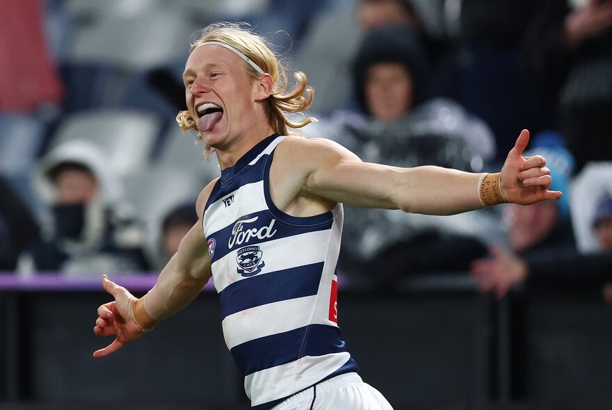 A Geelong AFL player celebrating on the field after having scored, his arms outstretched and tongue poking out, with blurred fans in the stadium in the background.