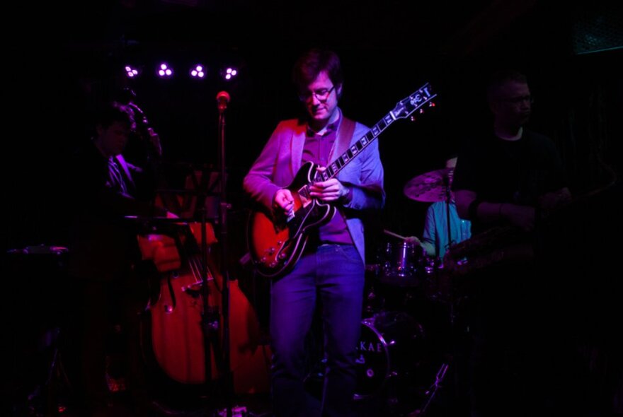 A performer playing guitar on stage in a dimly-lit venue.