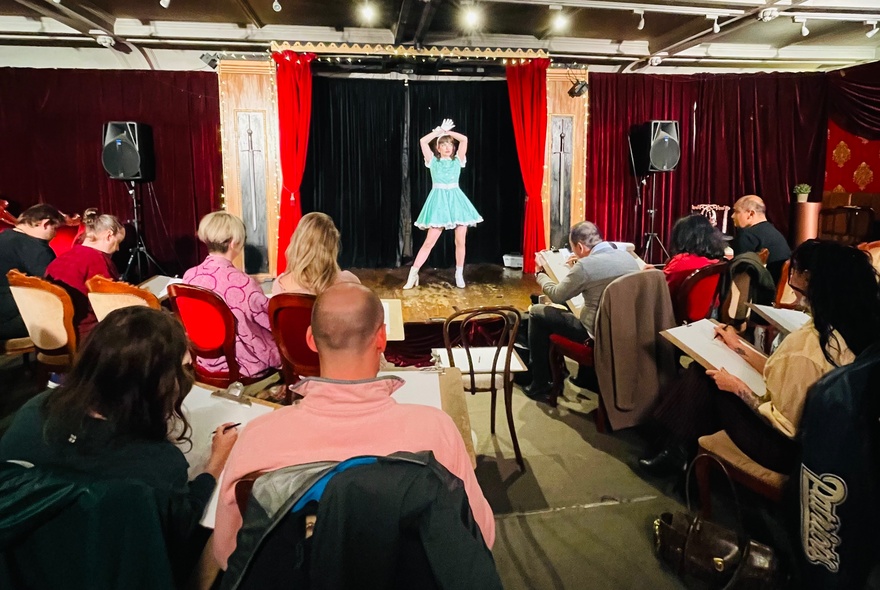 A burlesque performer in a blue dress poses with arms above her head in front of a small audience. 