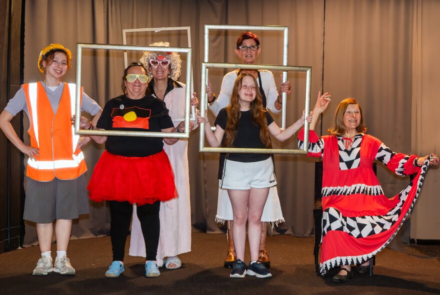A diverse group of performers pose on a stage while holding empty picture frames, in front of a stage curtain.