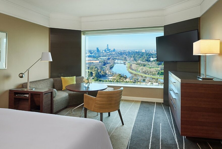 Hotel room with panoramic view of Melbourne and the Yarra River, with bed, sofa, table and cupboard.