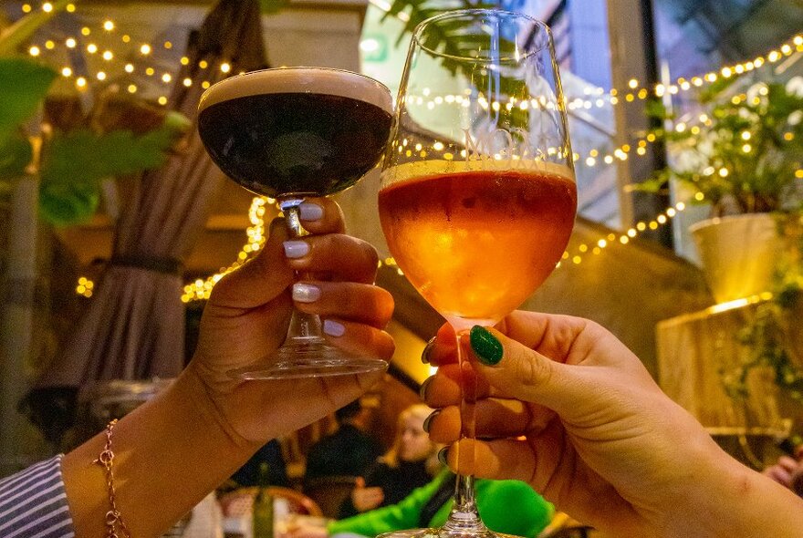 People holding up a wine and espresso martini in front of sparkly lights in an outdoor bar.