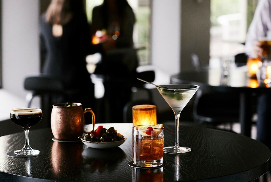 Candlelit bar table with cocktails, metal jug and bowl of olives.