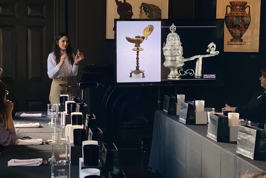 Presenter at a screen holding perfume in front of people seated at tables.