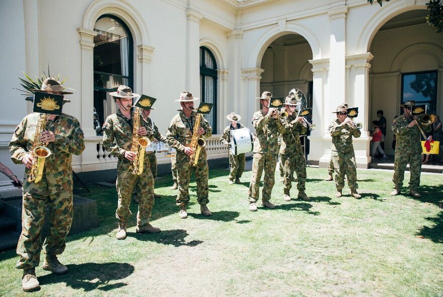 Soldiers playing brass instruments, wearing khaki uniforms, in a grassed sunny spot outside Government House.
