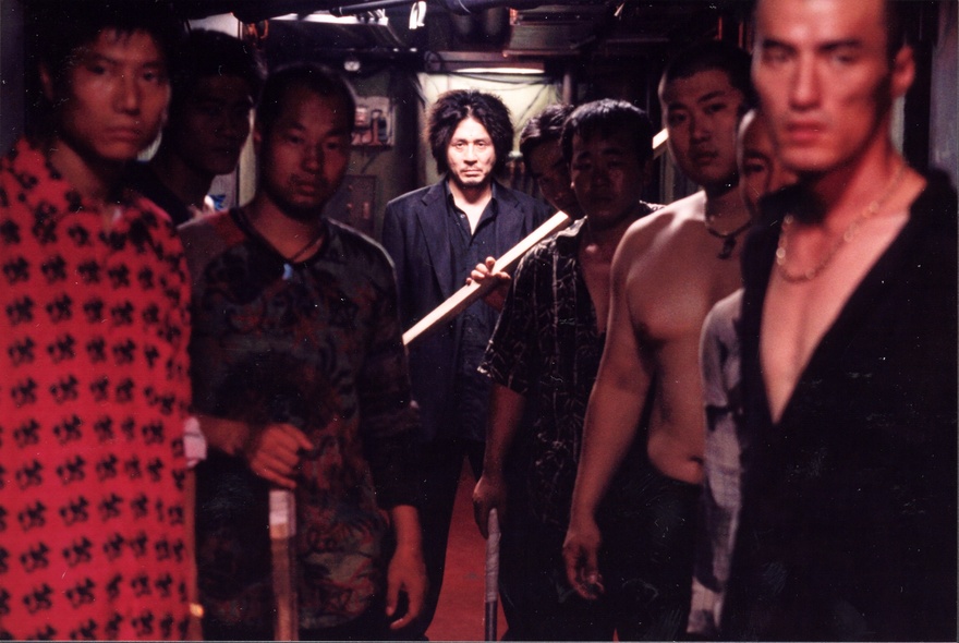 A still image from the film Oldboy, showing a man in a spotlight at the back of a group of dimly-lit men, holding a stick. 