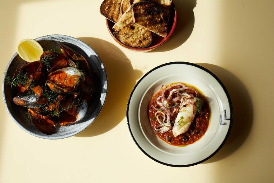 Three dishes of food on a table including grilled bread and a bowl of oysters with lemon.