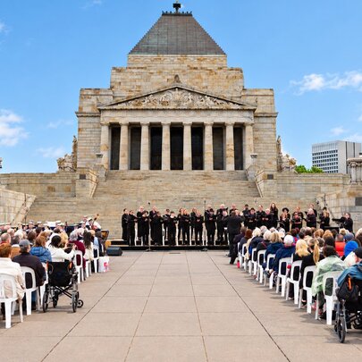 A commemorative gathering with a choir performing in front of the Shrine of Remembrance in Melbourne, and an audience seated on chairs in the forecourt.