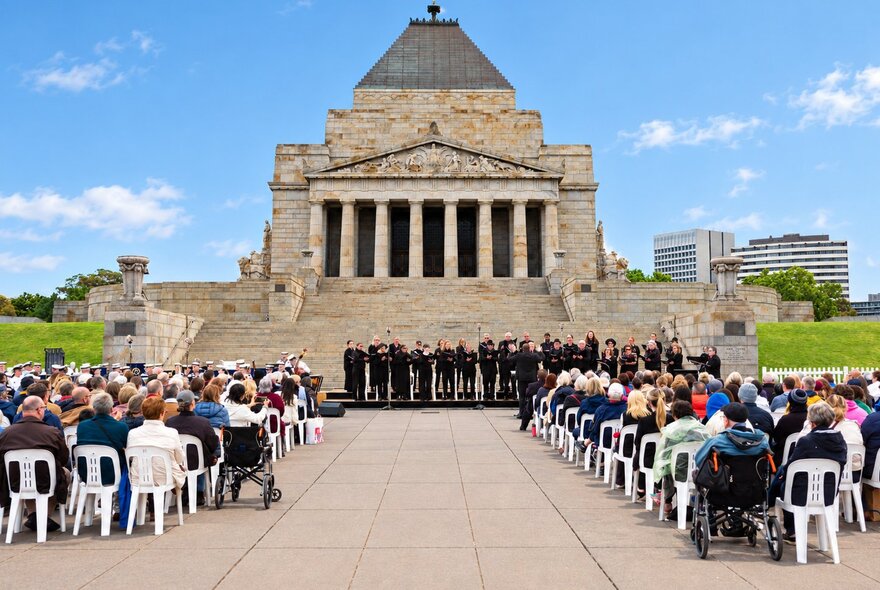 A commemorative gathering with a choir performing in front of the Shrine of Remembrance in Melbourne, and an audience seated on chairs in the forecourt.