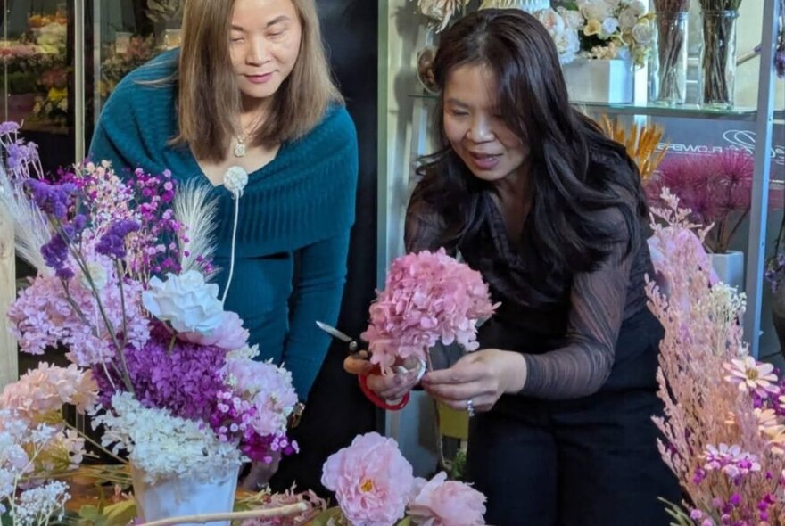 A woman cutting flower stems in a florist with another woman looking on.