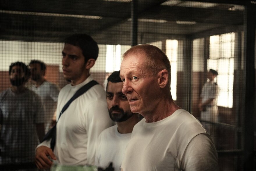 A still from the movie The Correspondent with actor Richard Roxburgh in a white t-shirt in a crowded room