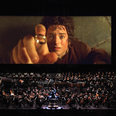 A full orchestra on a stage in front of a large cinema screen showing a still from the movie Lord of the Rings: The Fellowship of the Ring.