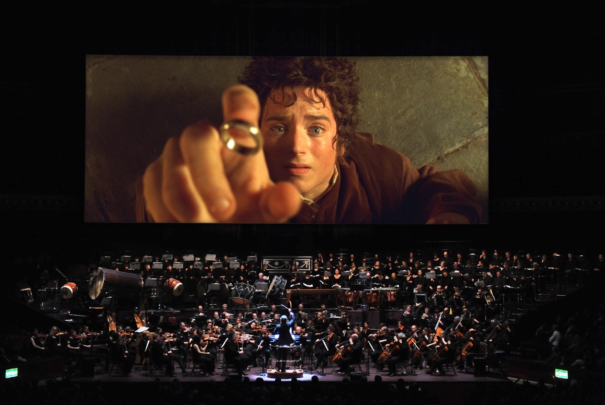 A full orchestra on a stage in front of a large cinema screen showing a still from the movie Lord of the Rings: The Fellowship of the Ring.