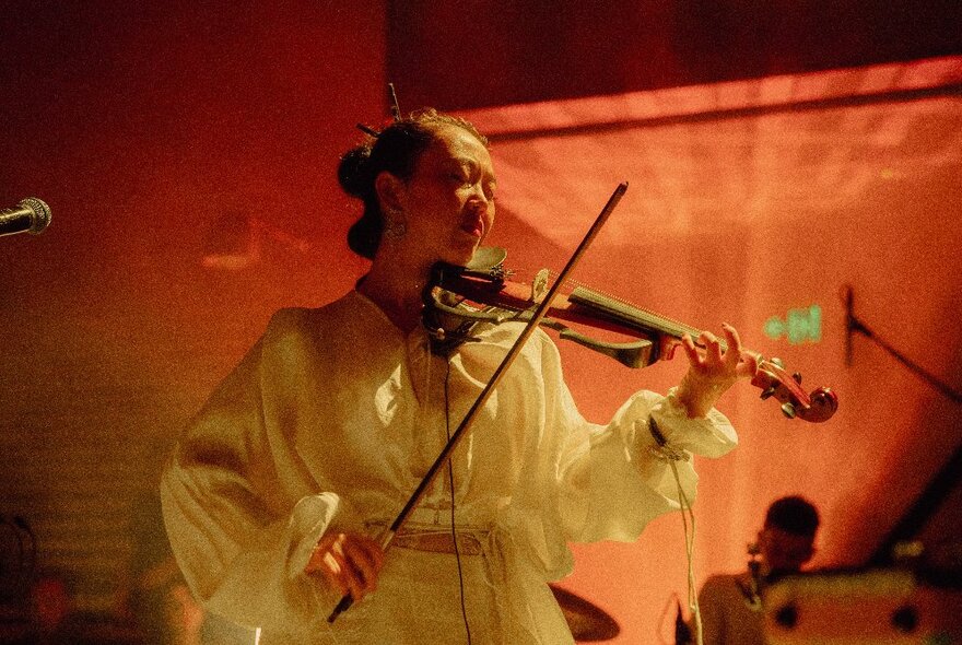 A woman in a white flowing dress playing a violin on a stage lit by orange coloured lights.