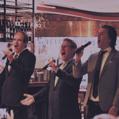 Three tenor singers singing into hand-held microphones in front of the bar in a restaurant setting.
