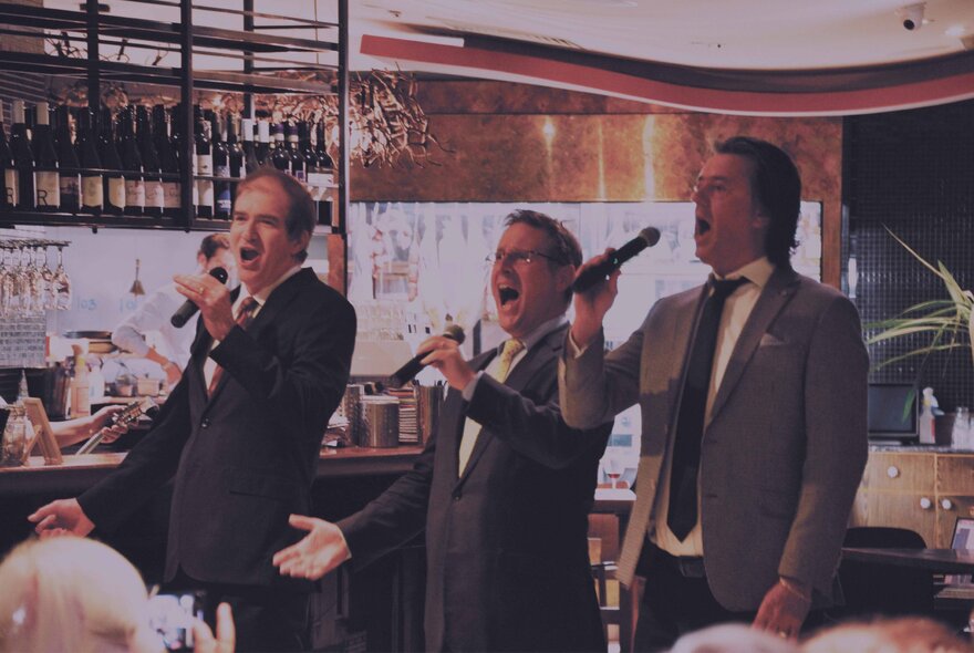 Three tenor singers singing into hand-held microphones in front of the bar in a restaurant setting.