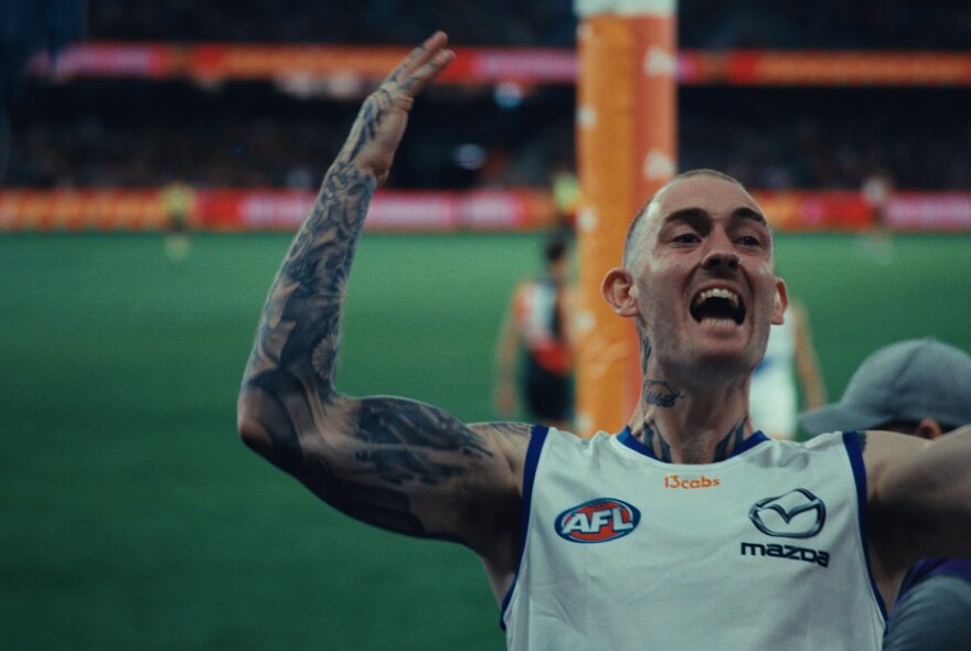 An Australian rules football player with a shaved head and a heavily tattooed arm celebrates enthusiastically on an AFL field.