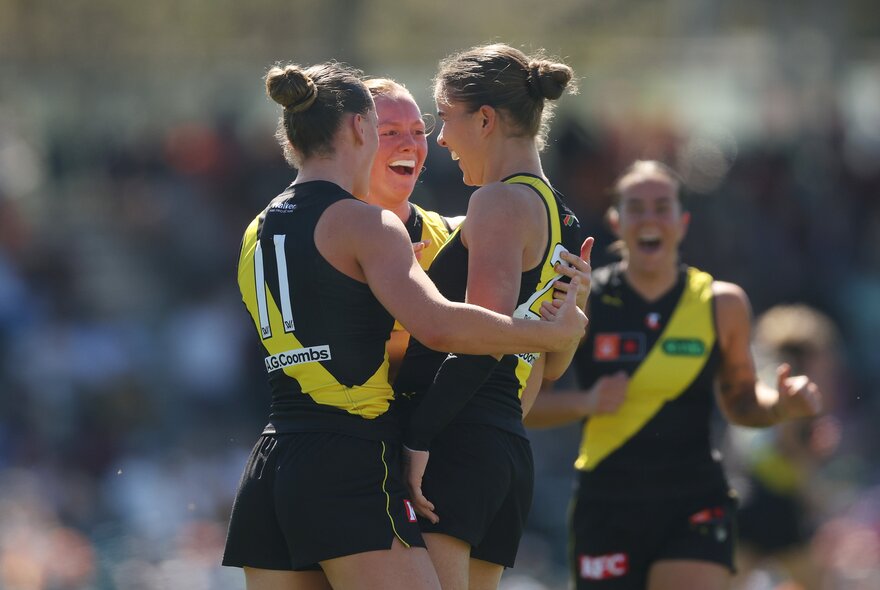 Three Richmond AFLW players celebrating on the field, coming together for a hug, with another in the background.