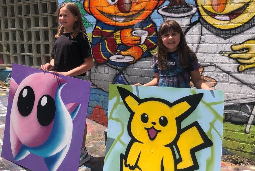 Kids holding Pokemon artworks outside, in front of a mural-covered brick wall.