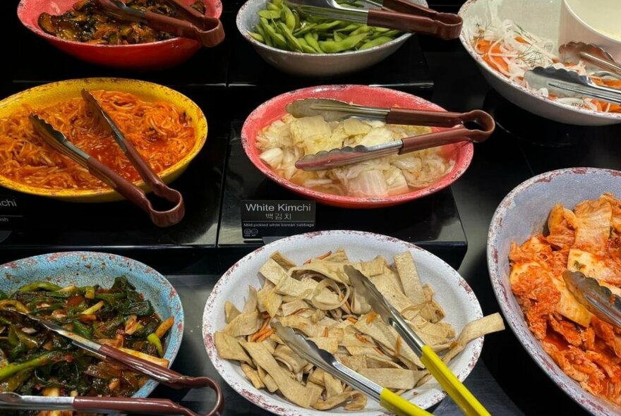 Dishes of Korean vegetables with tongs for serving.