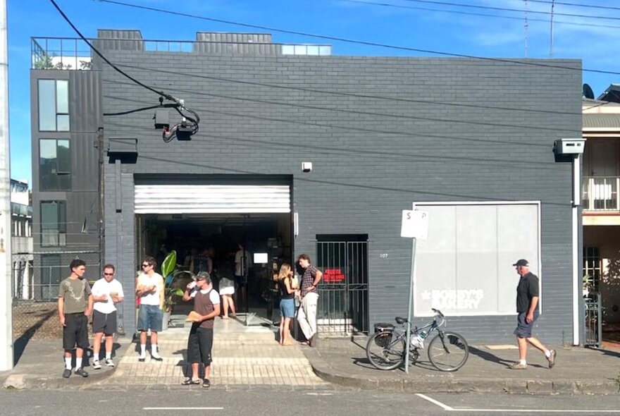 The roller door and grey wall exterior of Bobby's Bakery in a North Melbourne street, with people standing outside the front of the shop.