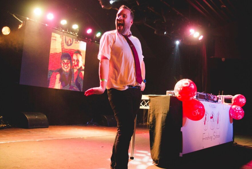 A man on stage with a projector screen behind him, and a table with black tablecloth and red balloons.