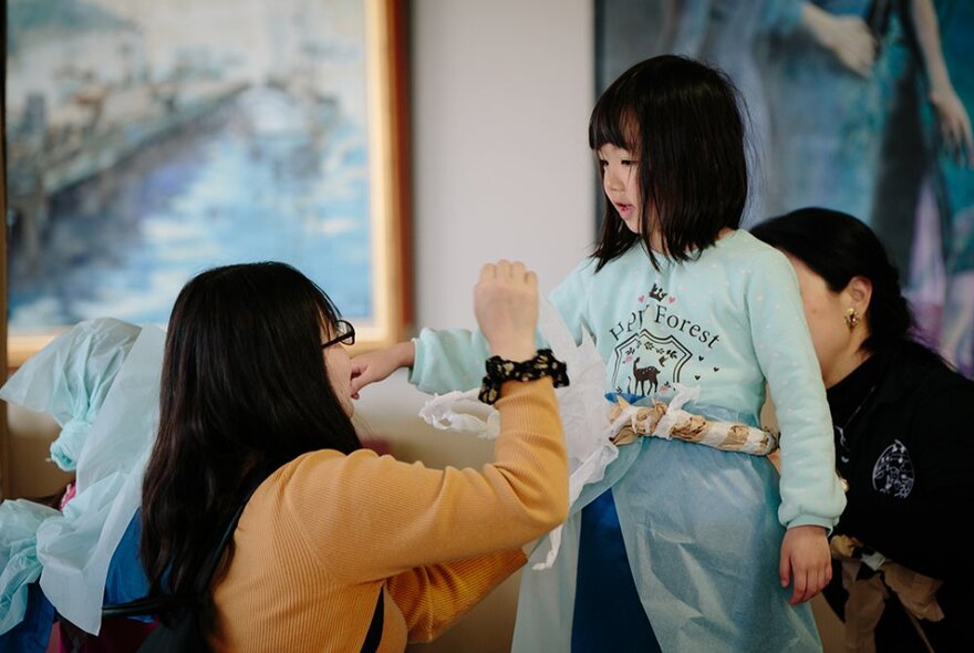 A small child looking down at her mother who is kneeling in front of her, blurred artworks on the wall behind them.