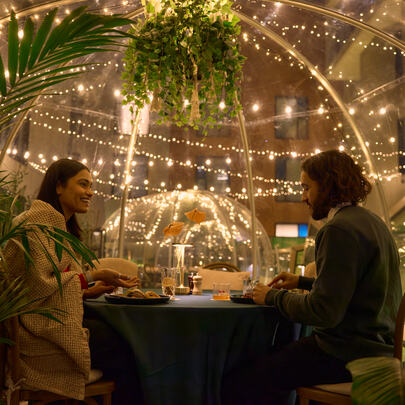 Two diners sit at a table under a dome decorated with fairy lights and plants