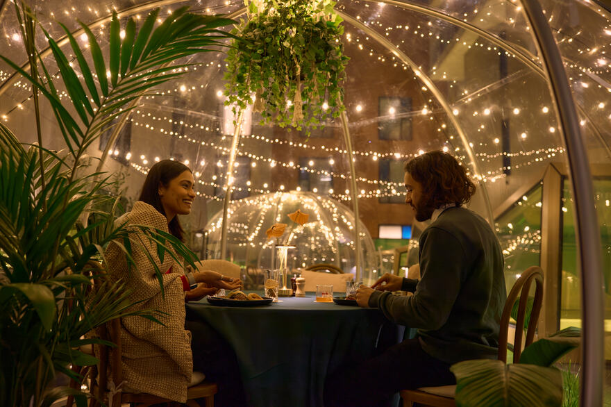 Two diners sit at a table under a dome decorated with fairy lights and plants