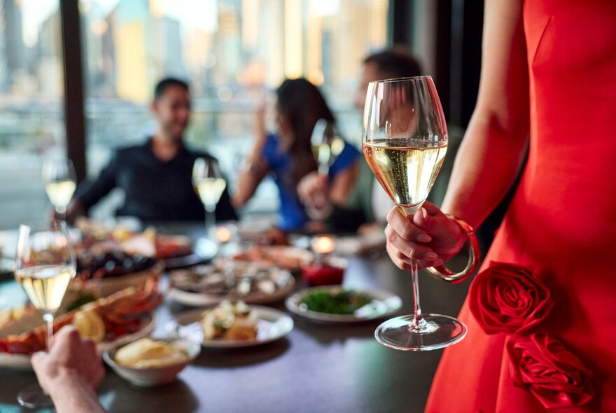 Woman in red gown holding a glass of champagne, diners at table in background with blurred city views.