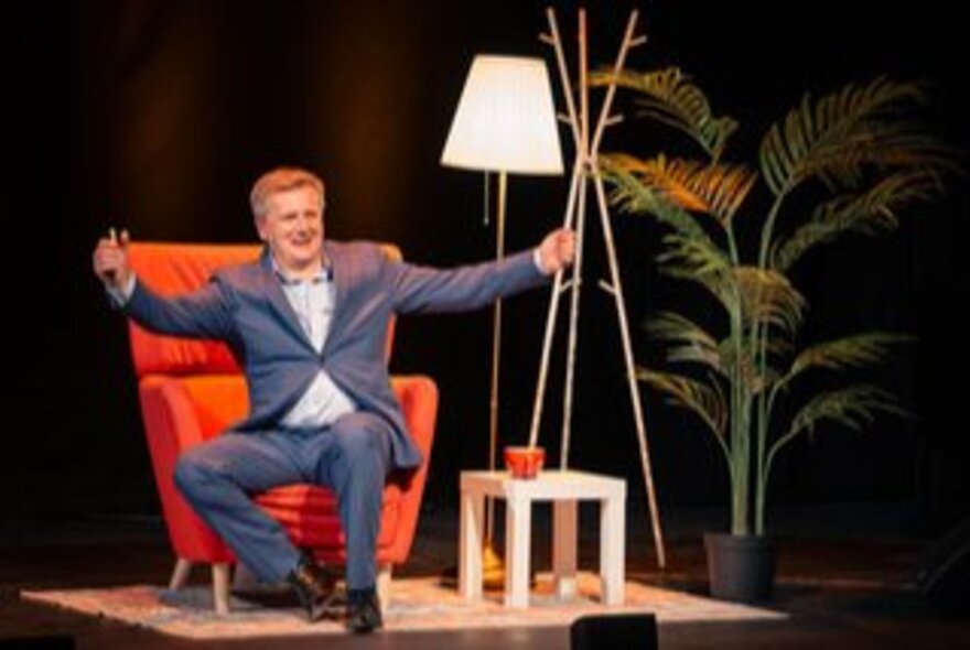 Aled Jones, wearing wearing a suit and seated in a large red chair on a stage, with a lamp and small coffee table next to him.