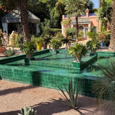 A bright green tiled geometric water fountain surrounded by exotic plants, palm trees, and gravel paths in the Majorelle Garden located in  Morocco.