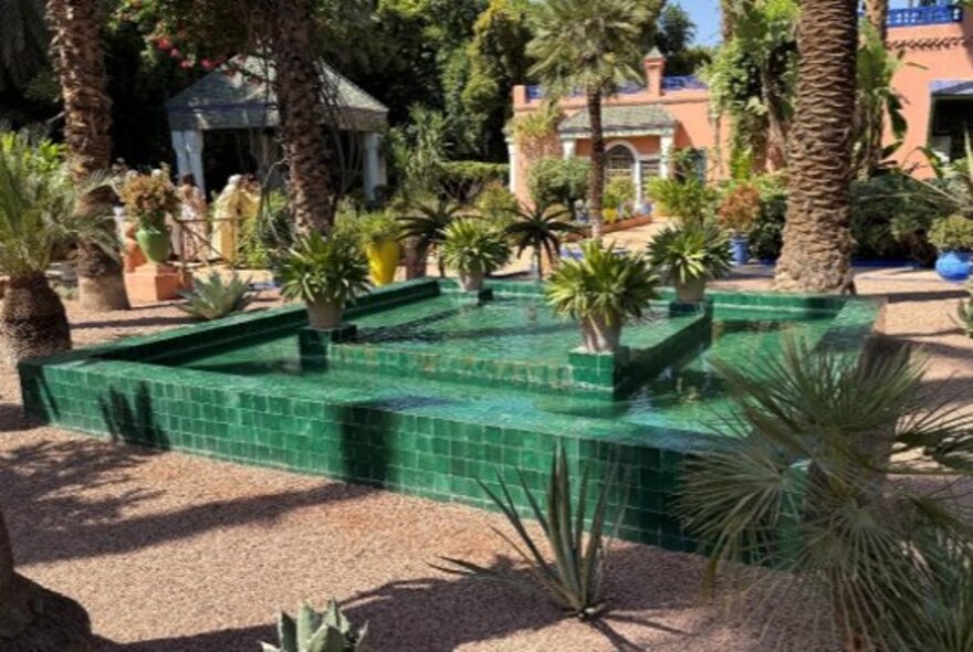 A bright green tiled geometric water fountain surrounded by exotic plants, palm trees, and gravel paths in the Majorelle Garden located in  Morocco.